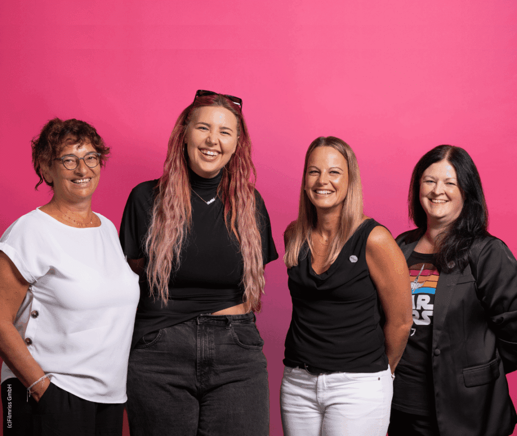 Die Künstlerin PÄM und die Geschäftsführerin, die stellvertretende Vorsitzende und Fundraiserin von MOKI NÖ stehen lächelnd vor einem pinken Fotohintergrund.